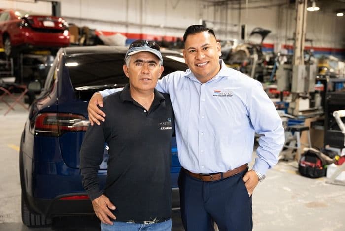 Two men smile in an auto shop, one in a cap and the other in a collared shirt.