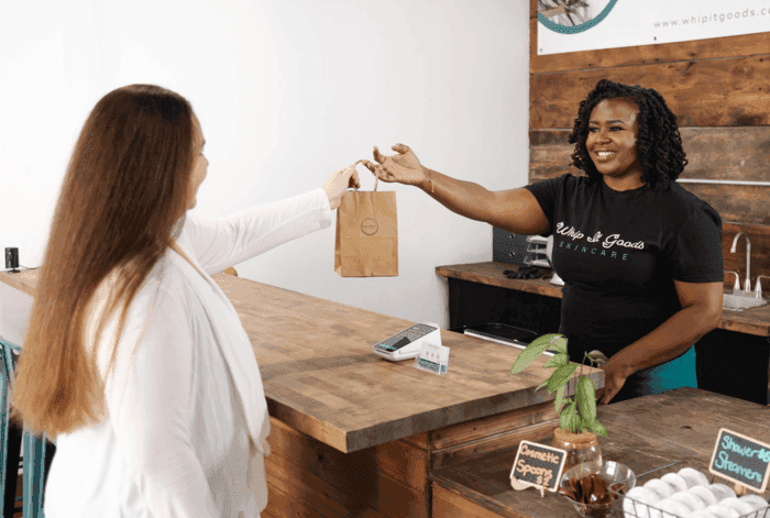 A woman hands a small brown paper bag to a customer over a rustic wooden counter.