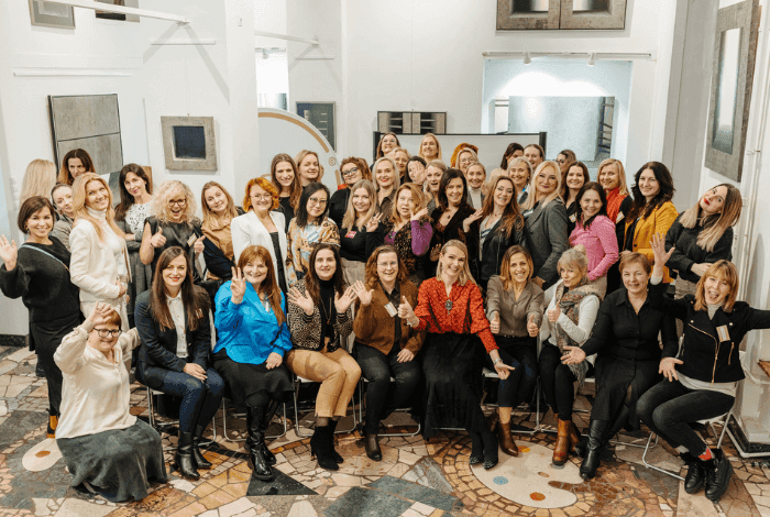 A large group of diverse women smiling and waving together in an indoor setting.