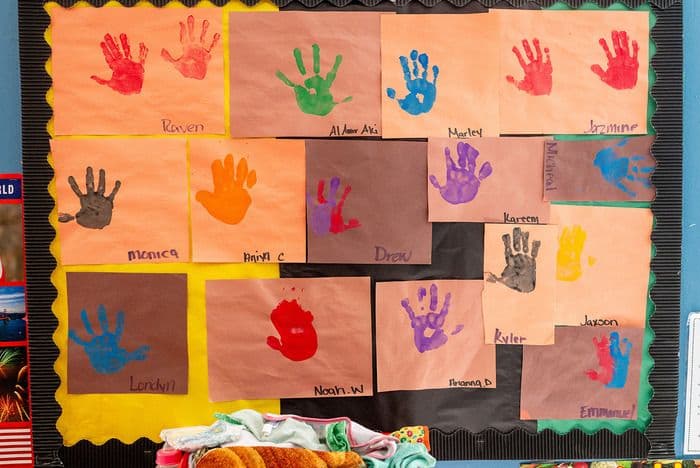A classroom bulletin board displays colorful handprints made by children on brown paper squares.