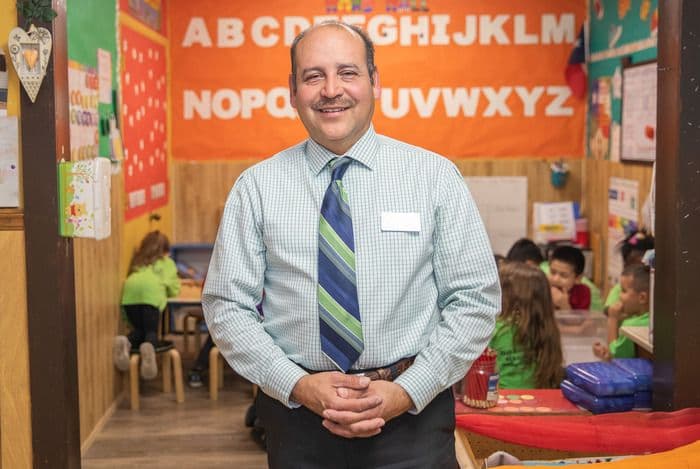 Smiling man in striped tie stands in a vibrant classroom with an alphabet banner.