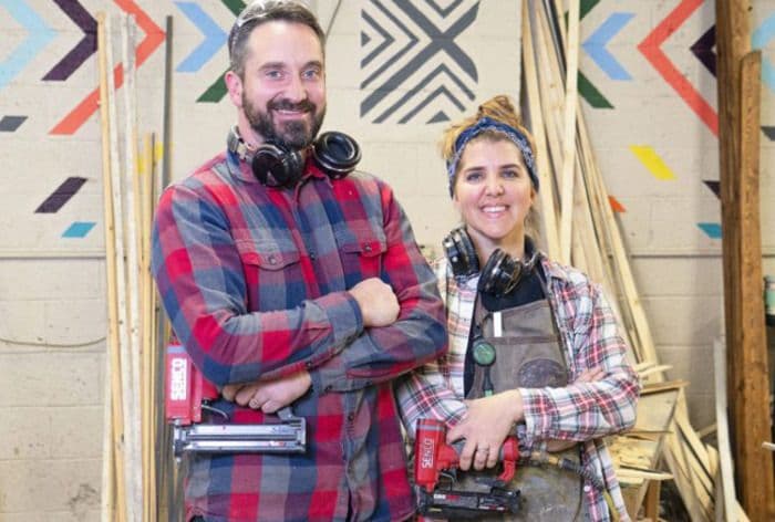 Smiling man and woman in plaid shirts standing in a workshop with tools.