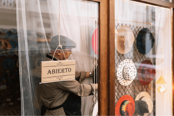 Man hangs an 'Abierto' sign in a shop window displaying various hats.