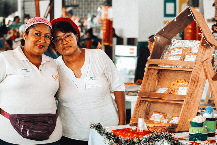 Two women smile next to a wooden market stall displaying packaged baked goods.