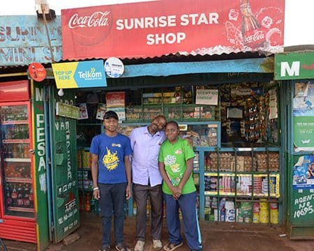 Three smiling men stand in front of a small 'Sunrise Star Shop' convenience store.