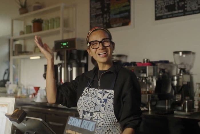 A smiling barista in glasses and apron waves from behind a coffee shop counter.
