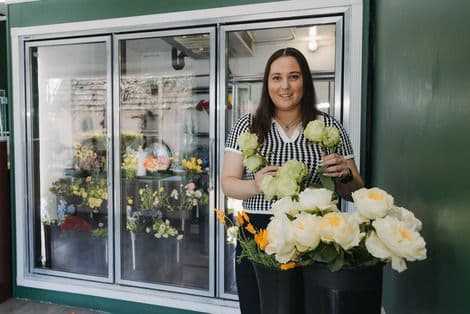 A smiling florist stands in front of a flower cooler, holding fresh light-colored roses.