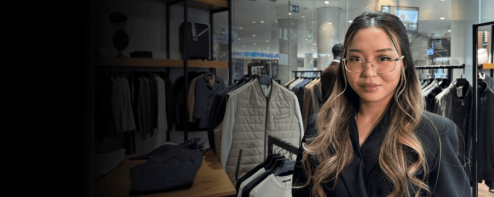 A woman with long wavy hair and glasses poses in a modern clothing store.