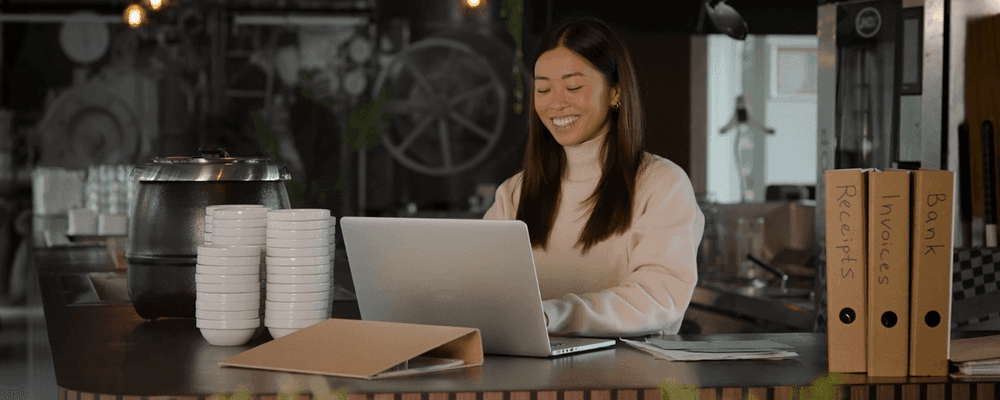 Smiling woman working on laptop at a cafe counter with files and cups.