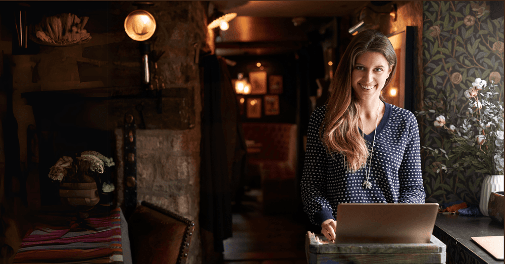 Smiling woman stands in a cozy pub, working on a laptop at a rustic counter.