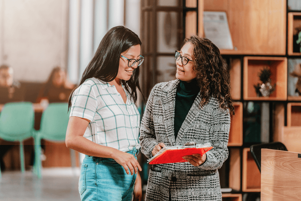 Two women with glasses discussing documents on a red clipboard in an office.