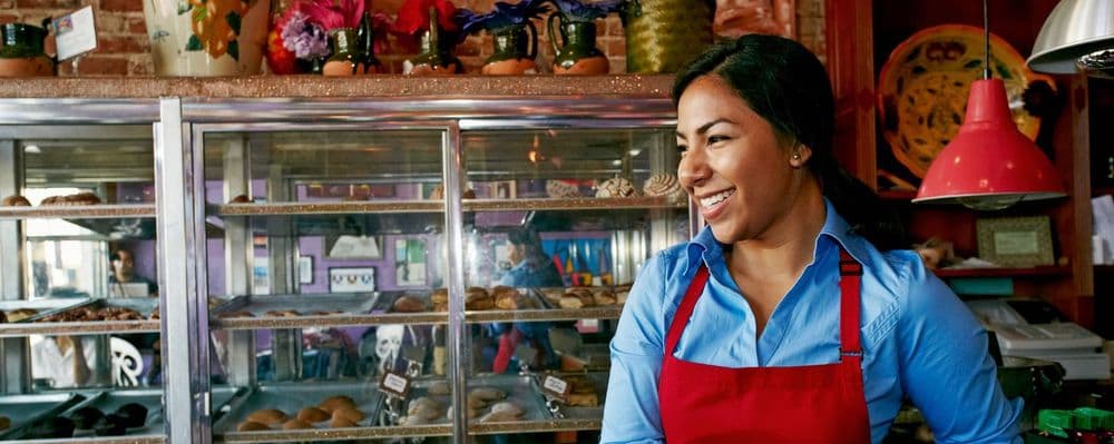 Smiling woman in blue shirt and red apron stands by bakery display cases.