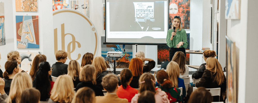 A woman in a green shirt presents to an audience in a bright room with art.