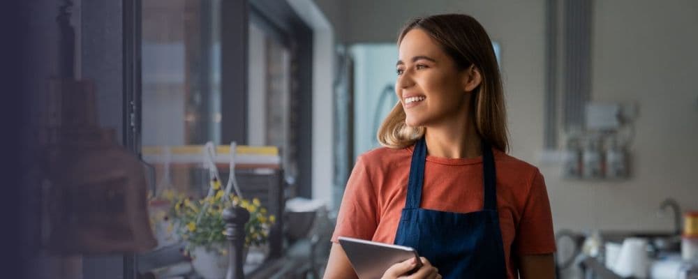 Smiling young woman in an apron holds a tablet, looking thoughtfully in a cafe setting.