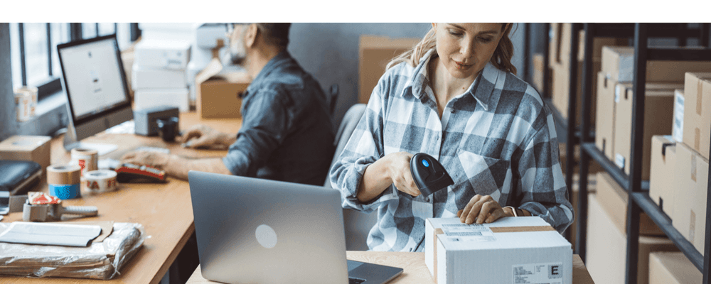 Woman scanning a package with a barcode scanner, man working on computer in background.
