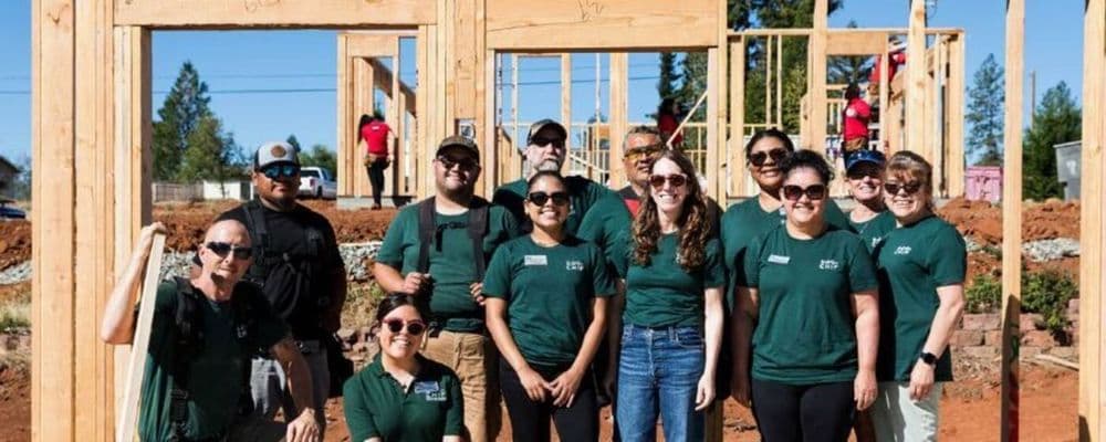 Diverse volunteers in green shirts pose proudly in front of a new house under construction.