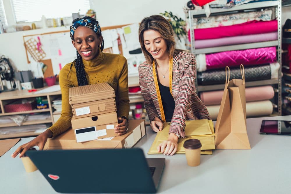 Two smiling women process orders on a laptop, packaging products in their design studio.