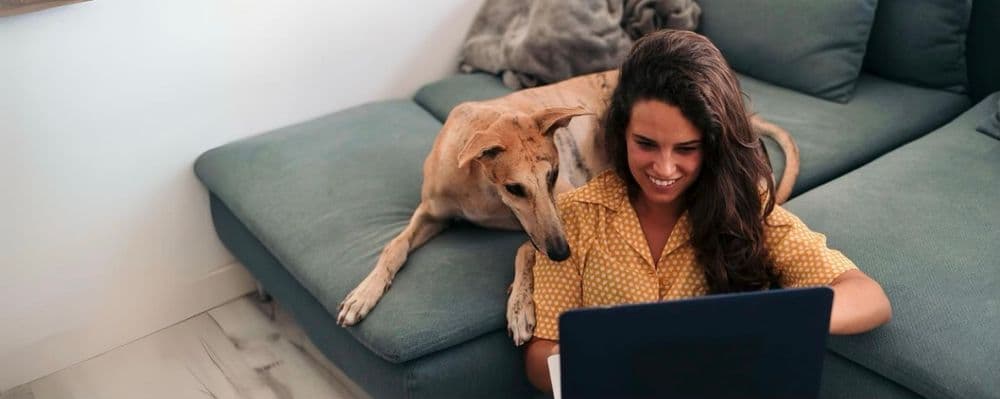 Woman on floor using laptop with her dog resting its head on her shoulder.