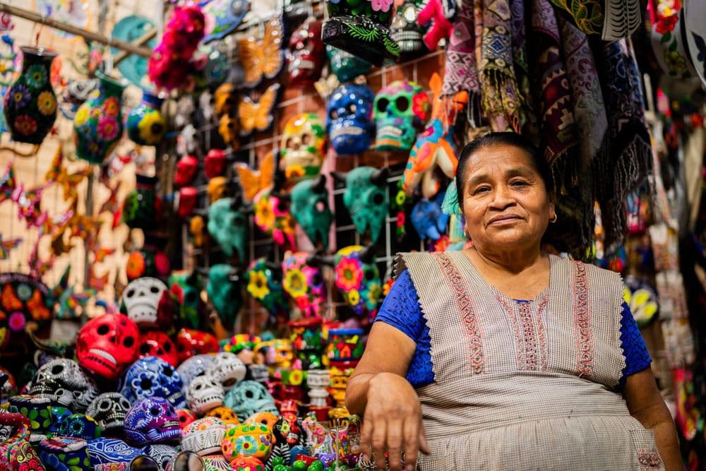 A woman in traditional dress stands proudly before a vibrant stall of Mexican skull crafts.