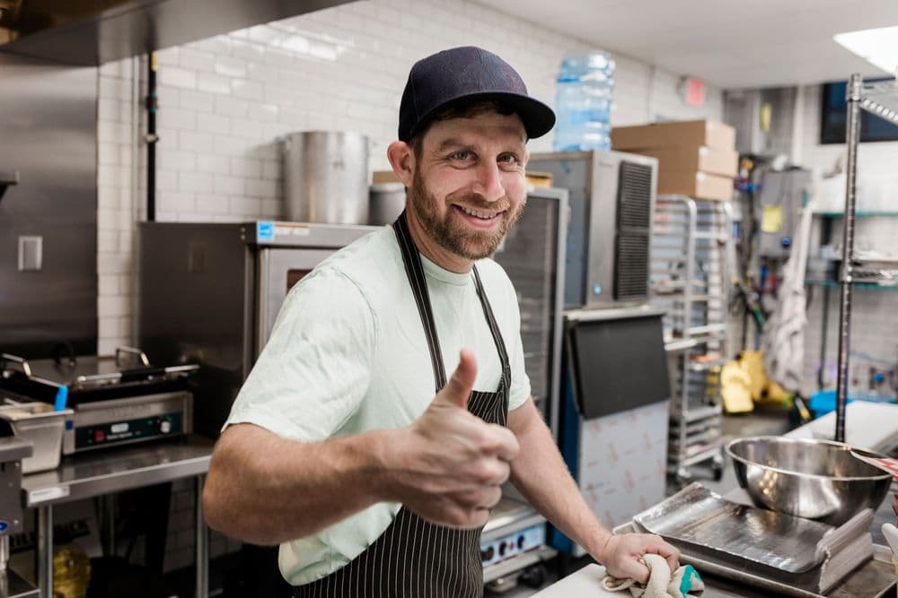 Smiling chef in a commercial kitchen wearing an apron and giving a thumbs-up.