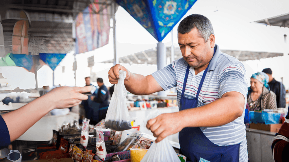 Market vendor in a striped shirt hands plastic bags of goods to a customer.