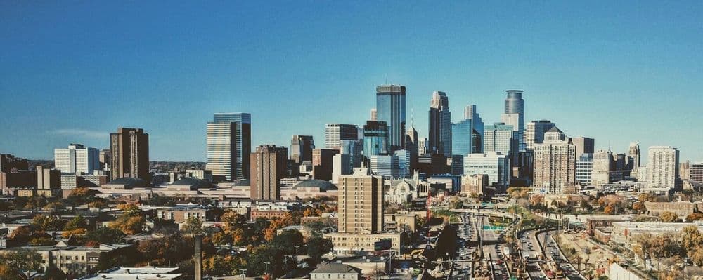 Panoramic city skyline with modern skyscrapers, autumn trees, and railway tracks under a clear blue sky.