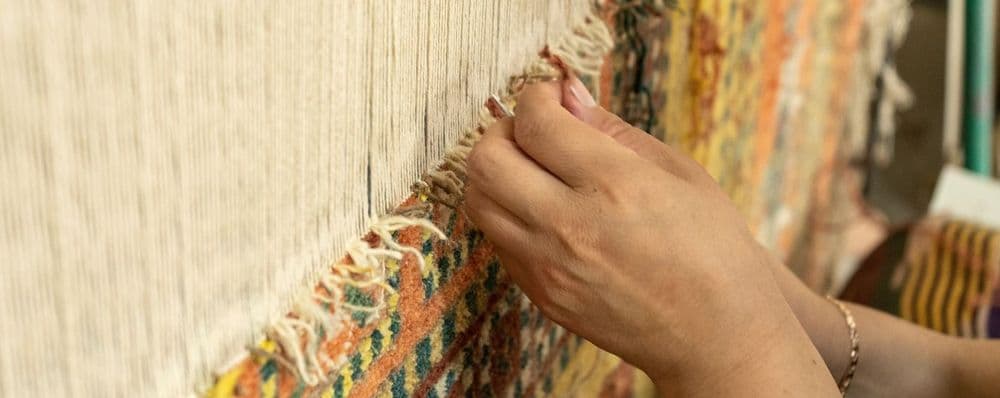 A person's hands meticulously weave colorful threads into a traditional rug on a loom.