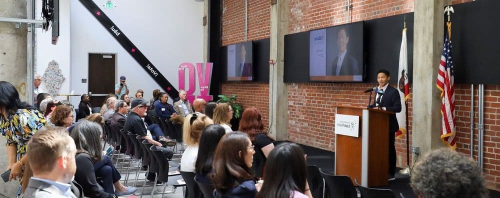 Man speaking at a podium to an audience in a large event room with flags.