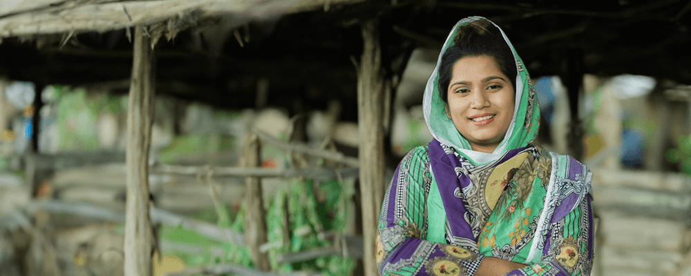 A smiling young woman in a colorful patterned headscarf and clothing stands outdoors.
