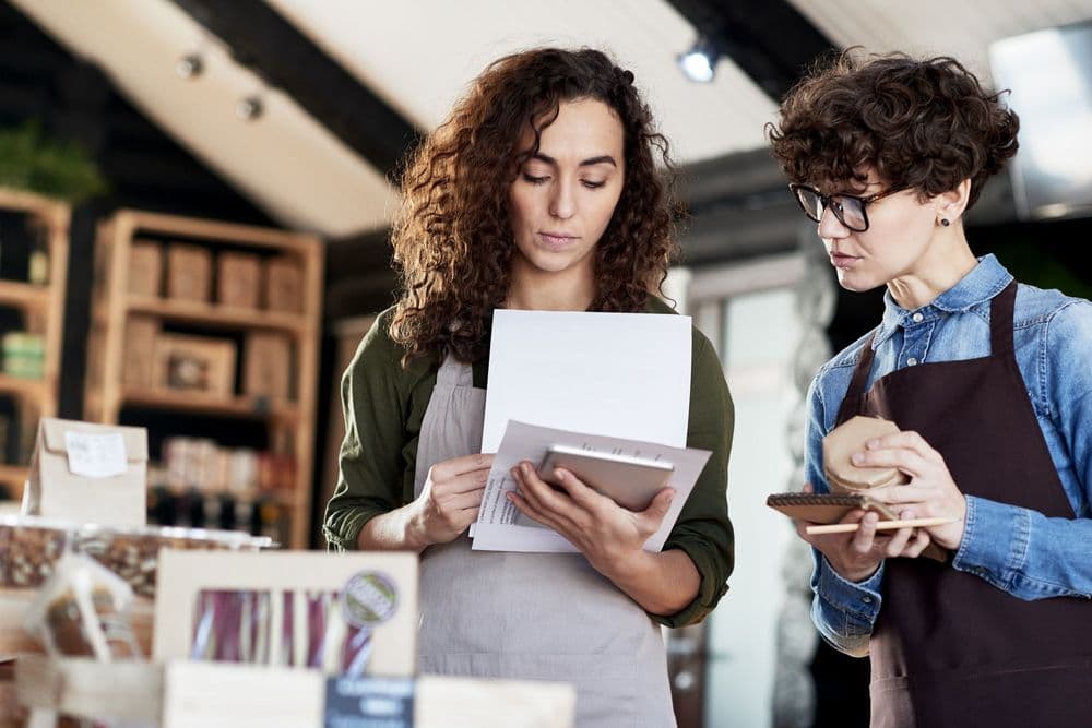 Two retail workers in aprons checking inventory, one holding papers, the other writing.
