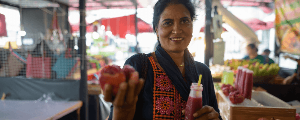 A smiling woman offers fresh fruit and a drink at an outdoor market.