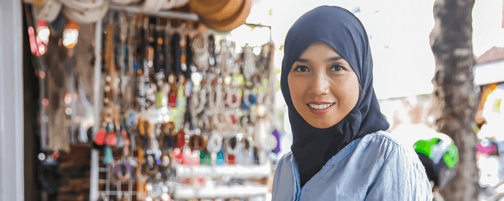 Smiling woman in hijab stands before a colorful market stall with various handmade goods.