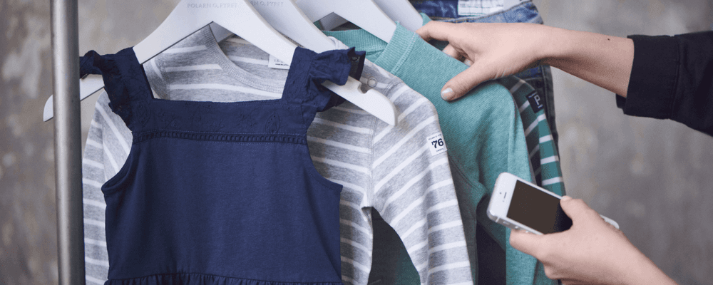 A person uses a smartphone to scan children's clothing hanging on a white rack.