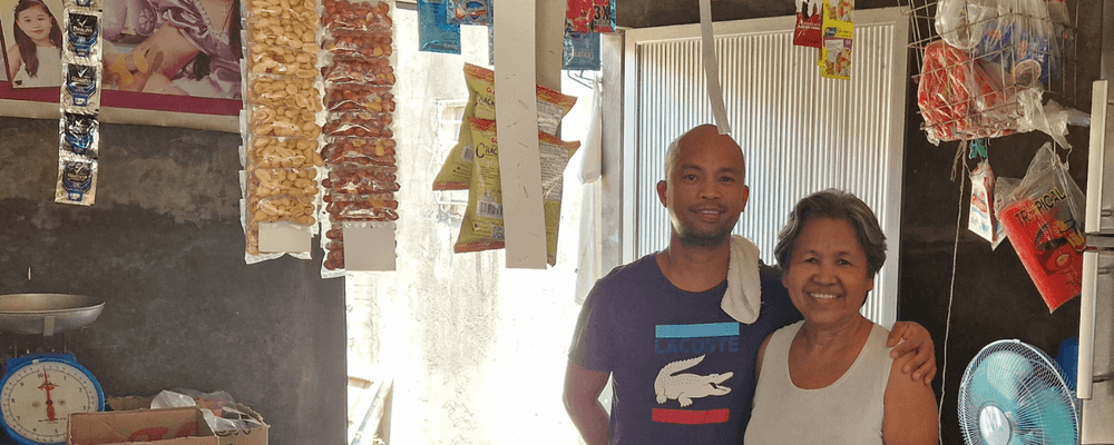 A smiling man and woman stand together in a small, cluttered market stall with various products.
