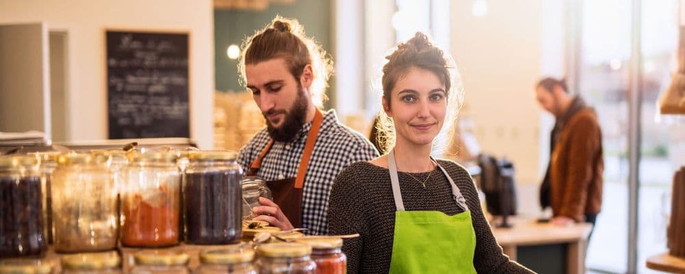 Young woman in a green apron smiles, while a man in an apron works in a store.