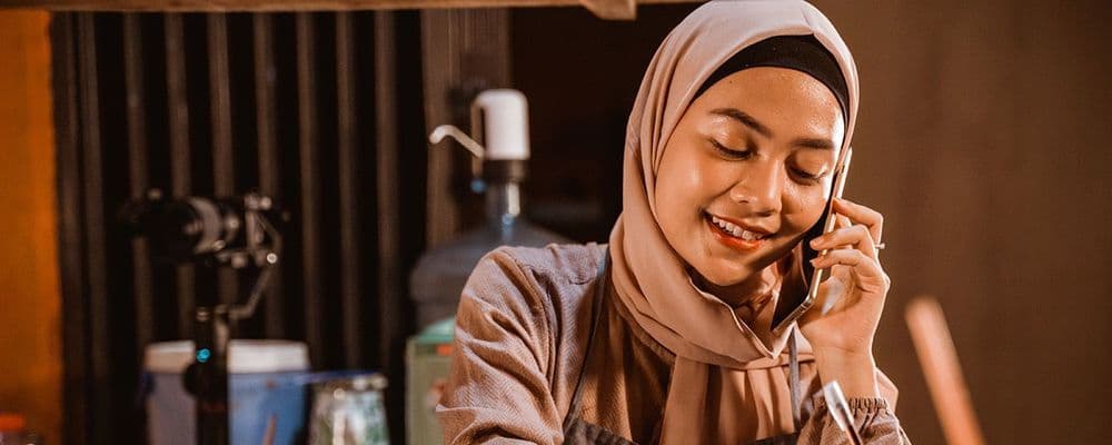 Smiling woman in hijab talks on a smartphone, looking down with a pleasant expression.