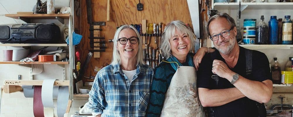 Three smiling craftspeople, two women and one man, stand in their busy workshop.