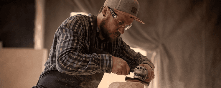 A craftsman wearing safety glasses and a cap sands a wooden object with a tool.