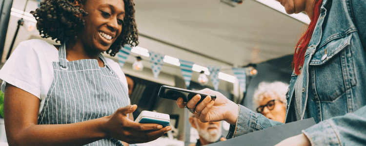 Smiling vendor accepts contactless payment from customer using smartphone at an outdoor stall.