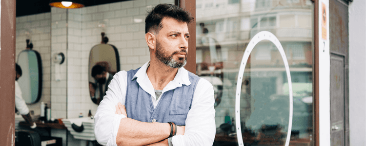 Bearded man, a barber or shop owner, stands proudly in his barbershop doorway.