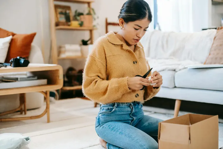 Woman in yellow cardigan kneeling, opening a cardboard box with scissors in her living room.