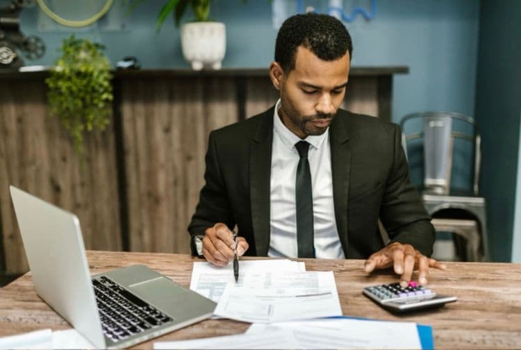 A man in a suit works at a desk, reviewing papers and using a calculator.