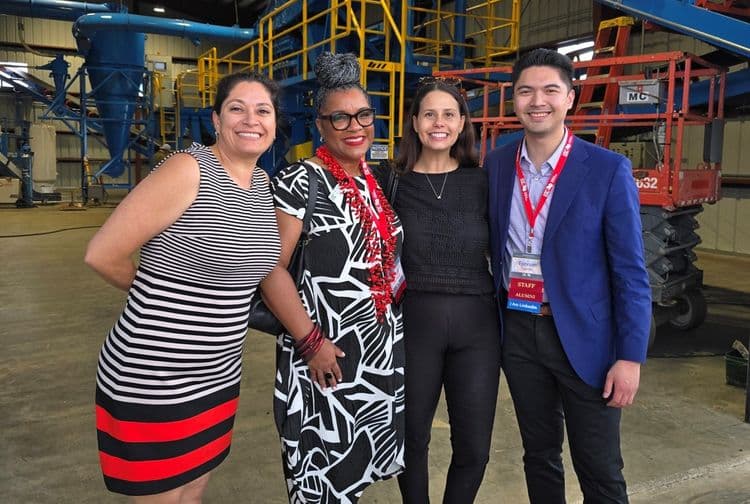 Four diverse professionals smile confidently in front of industrial machinery.
