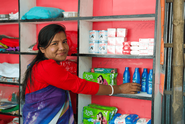 Woman in sari reaching for products on a red shelf in a small shop, smiling.