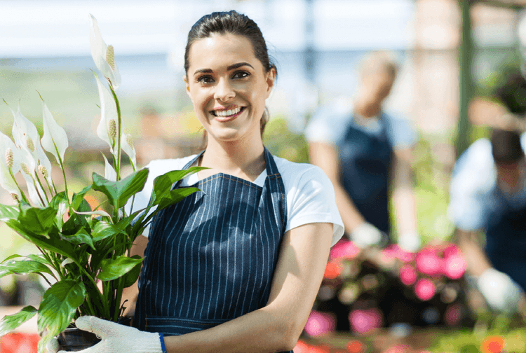 Smiling woman in apron and gloves holding a potted peace lily plant in a nursery.