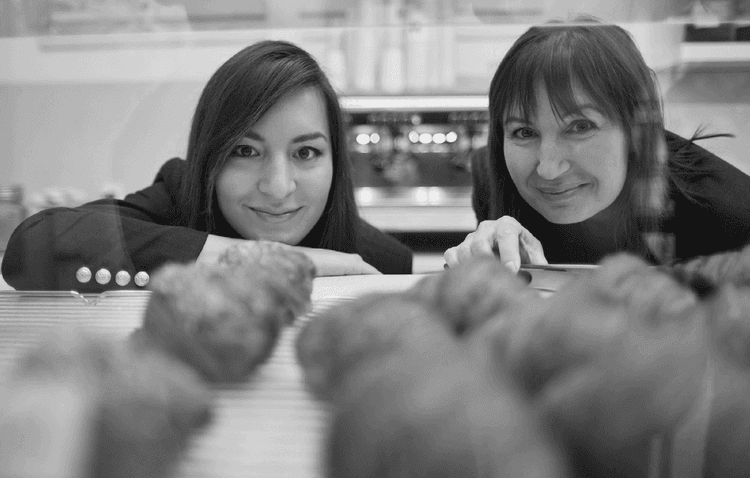 Two smiling women, likely business partners, look over a counter from a cafe.