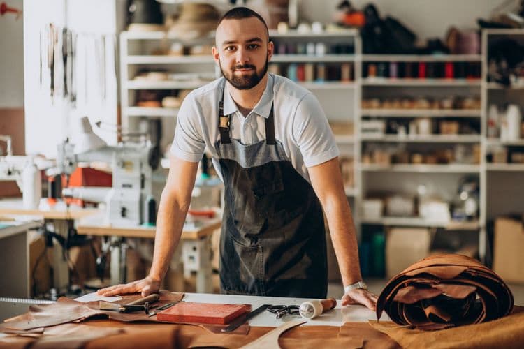 Man in an apron stands proudly in his leathercraft workshop, surrounded by materials.