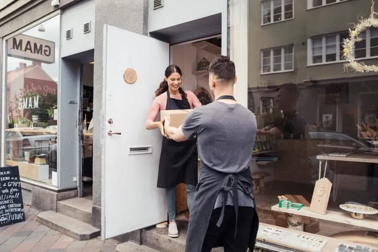 A woman in an apron happily accepts a package from a delivery driver outside a shop.