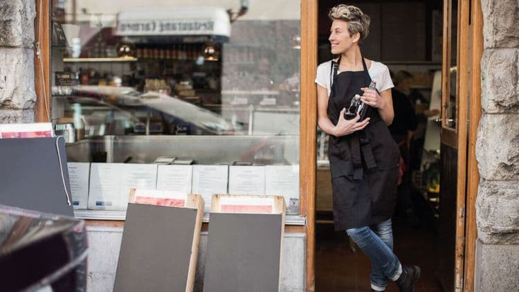 A smiling female business owner in an apron stands in her shop doorway.