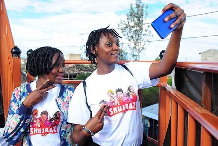 Two young women in matching Shujaaz t-shirts take a selfie with peace signs.